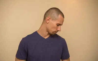 Hombre con un corte de pelo buzz cut, mirando hacia abajo con expresión pensativa.
