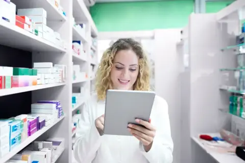 Farmacéutica sonriente revisa inventario en su tablet, rodeada de estantes llenos de productos de atida mifarma.
