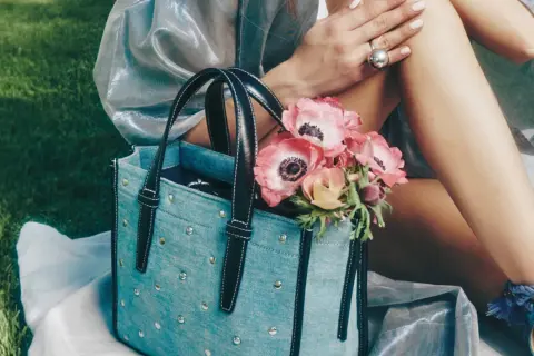 Un bolso de mezclilla azul con tachuelas y asas negras, adornado con un ramo de anémonas rosadas.
