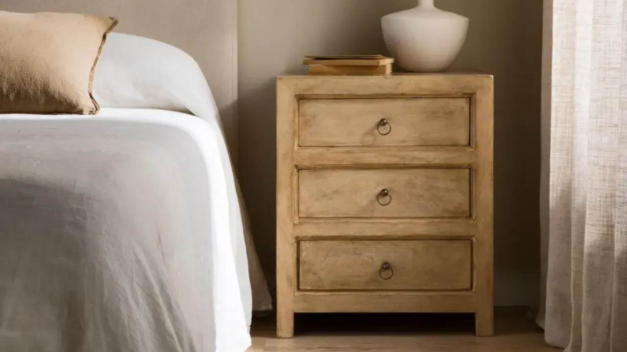 Mesita de noche de madera clara con tres cajones, parte de un dormitorio kenay home. Un jarrón blanco y libros decoran la superficie.