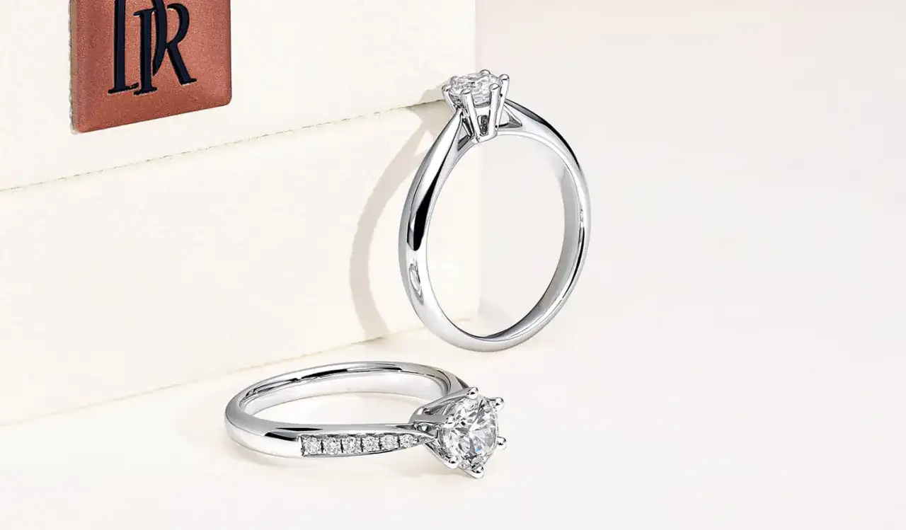 Dos anillos de compromiso de oro blanco con diamantes, uno con un solitario y otro con una banda de diamantes, junto a un logo de Darry Ring.