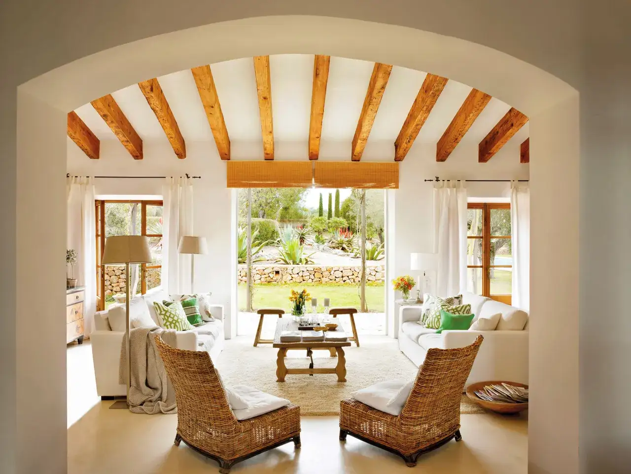 Salón luminoso con vigas de madera, sofás blancos y vistas a un jardín mediterráneo. Un rincón de maison du monde que invita a la relajación.