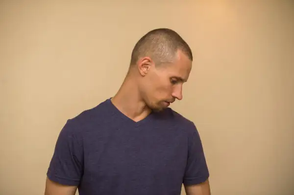 Hombre con un corte de pelo buzz cut, mirando hacia abajo con expresión pensativa.